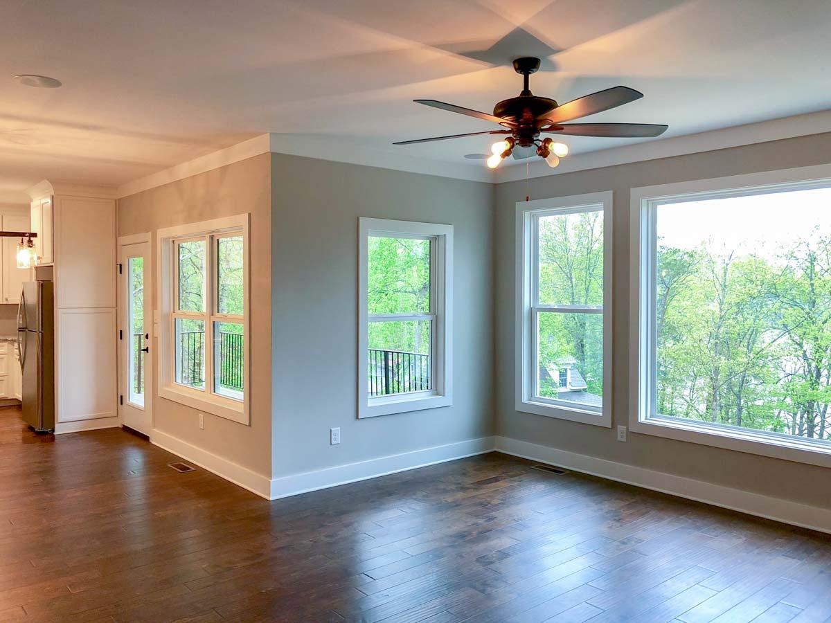 Interior view of a room with large windows overlooking trees, a ceiling fan, and a partial view of a kitchen with white cabinetry.