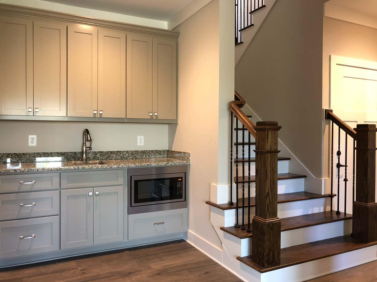 Interior view of a wet bar with gray cabinets, granite countertop, sink, microwave, and a wooden staircase with black spindles.