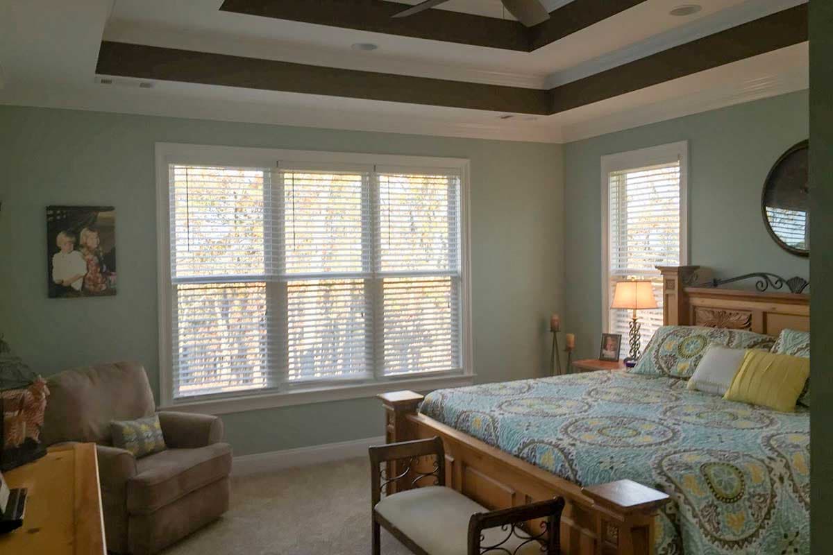 Bedroom interior with tray ceiling featuring dark beams, a large window with blinds, and a wooden bed.