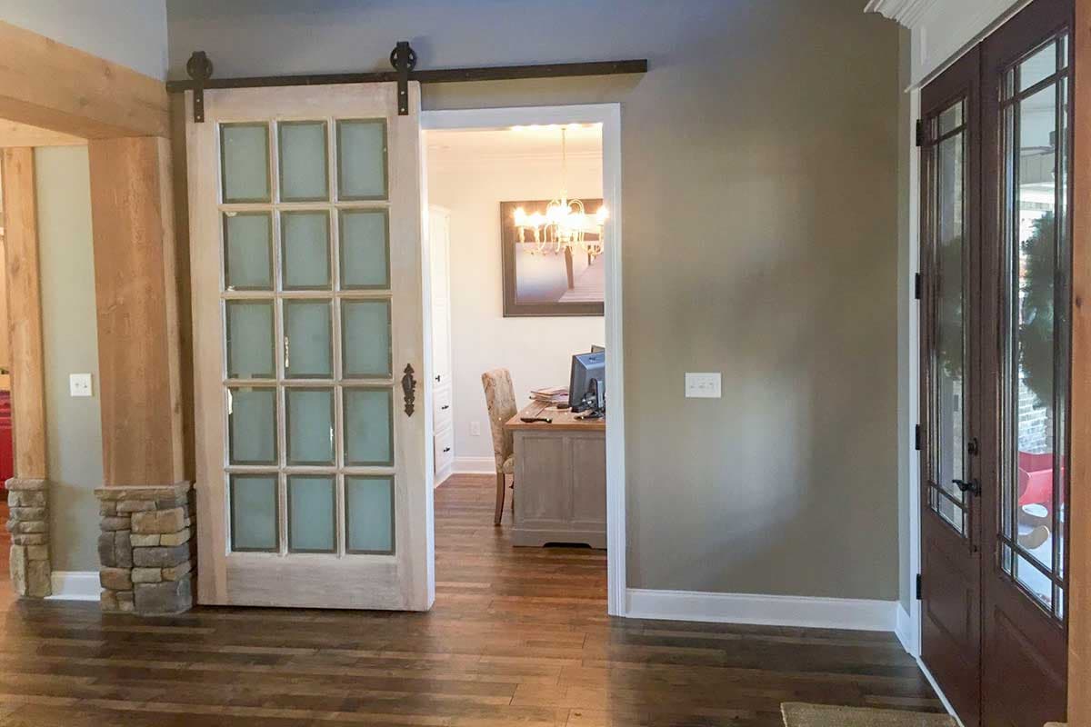Interior view shows a light wood sliding barn door with frosted glass panes opening to an office with a desk and chair.