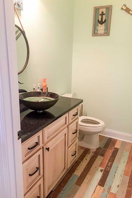 Powder room with a light wood vanity, dark countertop, vessel sink, toilet, and multi-colored plank flooring.