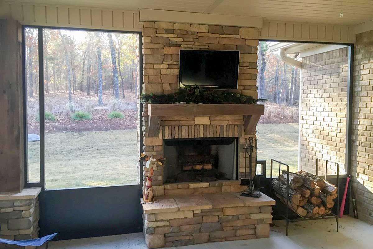Stone fireplace with timber mantle, television, and wood storage on a screened porch with large windows overlooking a wooded yard.