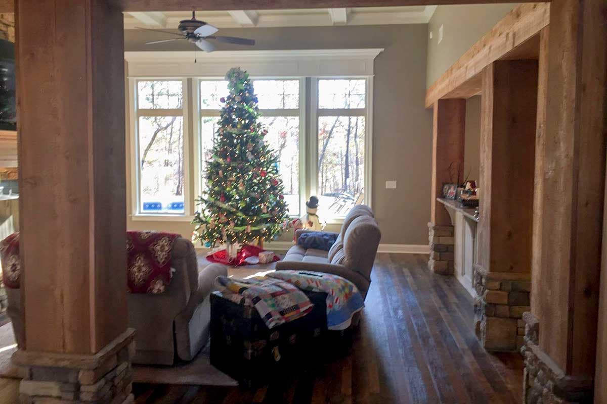 Interior view of living room with large windows, coffered ceiling, wood beams, stone pillar accents, and Christmas tree.