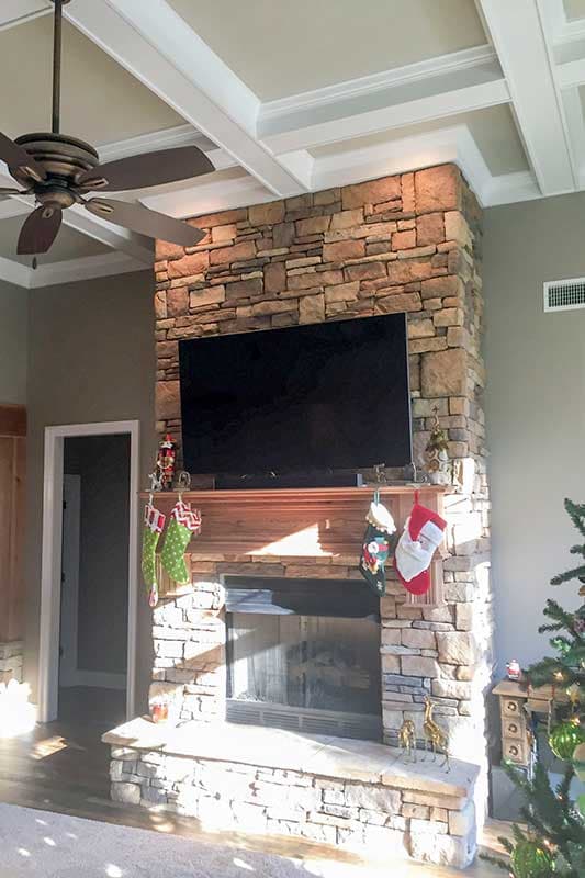 Great room fireplace with stone surround, wood mantel, coffered ceiling with fan, and mounted TV.