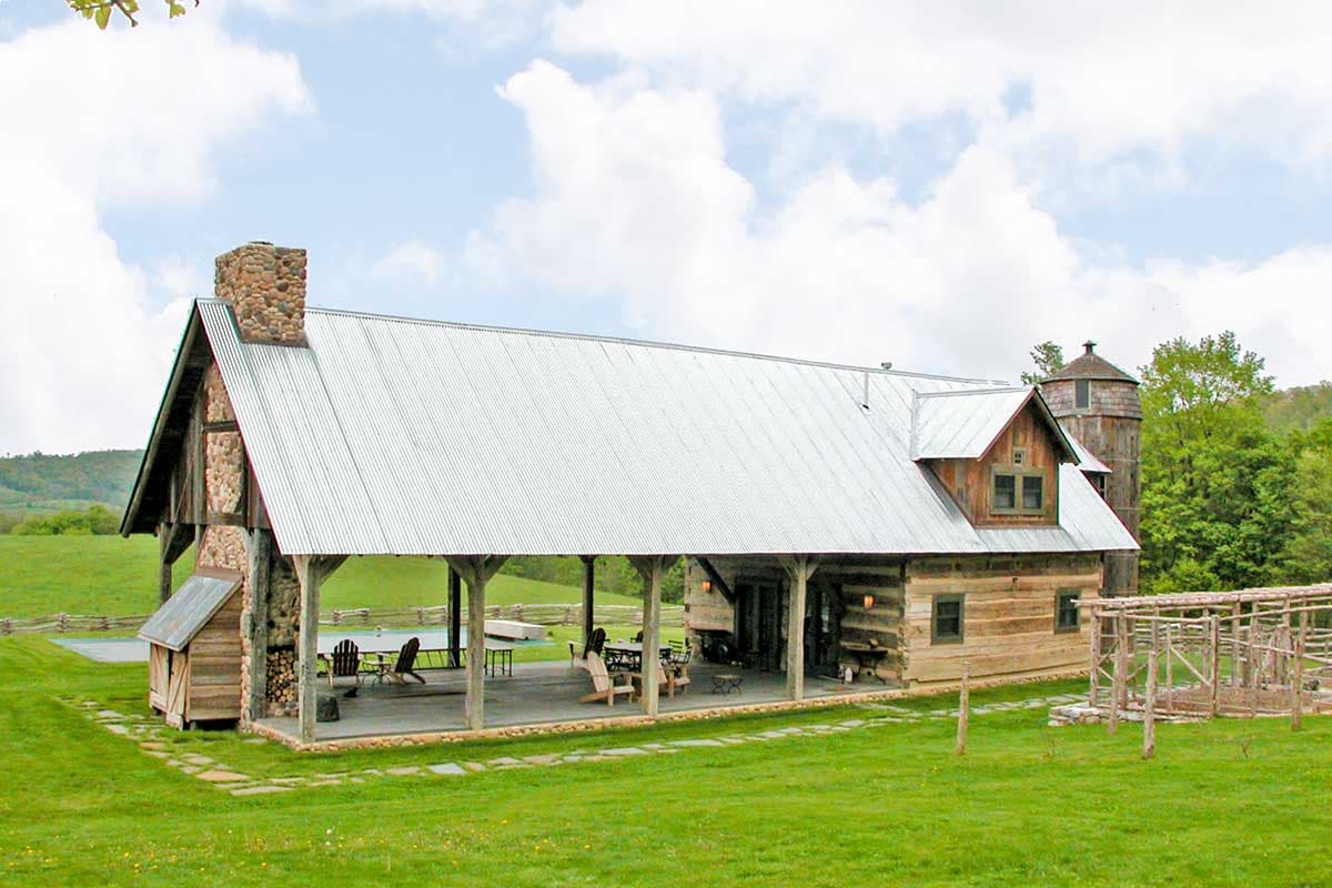 Log cabin house plan exterior with large covered porch, metal roof, stone chimney, dormers, and silo.
