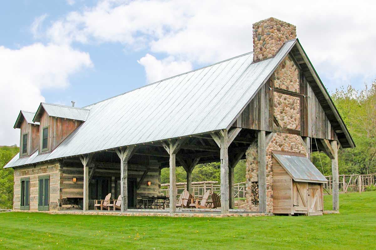 Log home exterior with large covered porch, stone chimney and metal roof. Features dormers and wood siding.