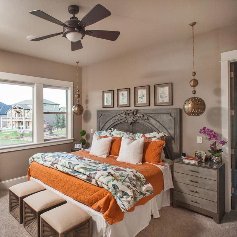 Interior view of a bedroom featuring a large bed with an ornate headboard, orange bedding, and large windows overlooking houses.