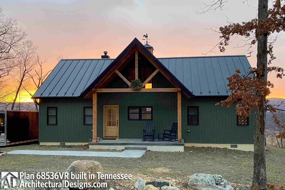 Modern cabin exterior with a steeply pitched metal roof, exposed timber framing, covered front porch with rocking chairs, and dark green siding.