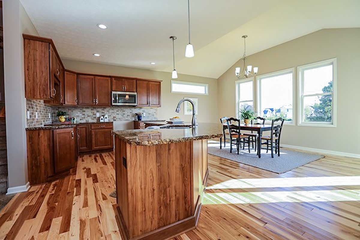 Open-concept kitchen with wood cabinets, granite island, dining area, vaulted ceiling, and large windows.