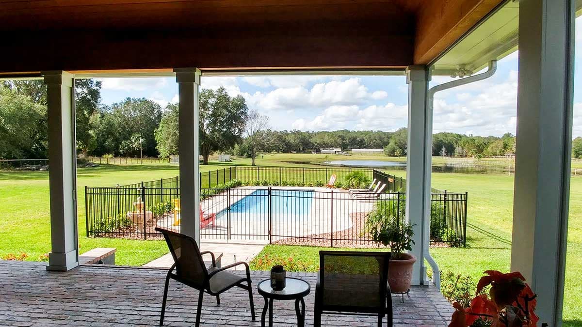 Covered porch with columns overlooking a fenced pool, lawn, and pond.