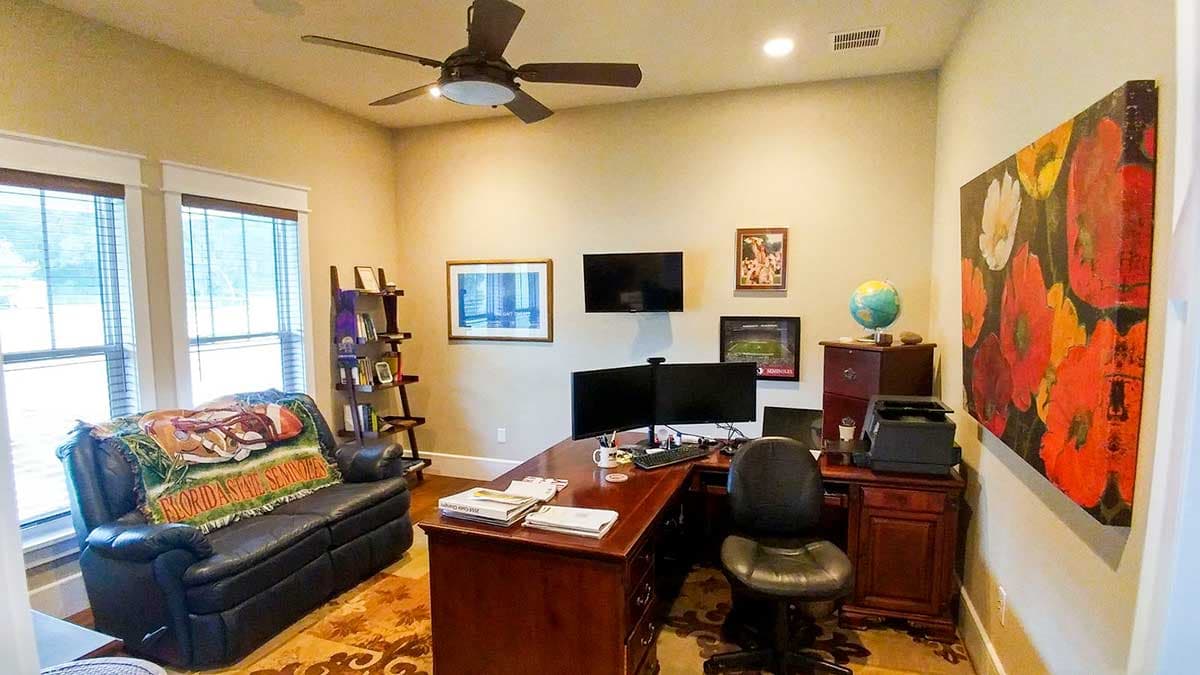 Home office with dark leather sofa, wooden desk with dual monitors, ceiling fan, and floral art.