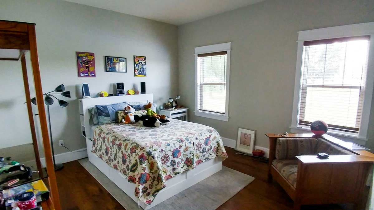 Bedroom with white bed, two windows with blinds, and wooden floor. Artwork and stuffed animals visible.