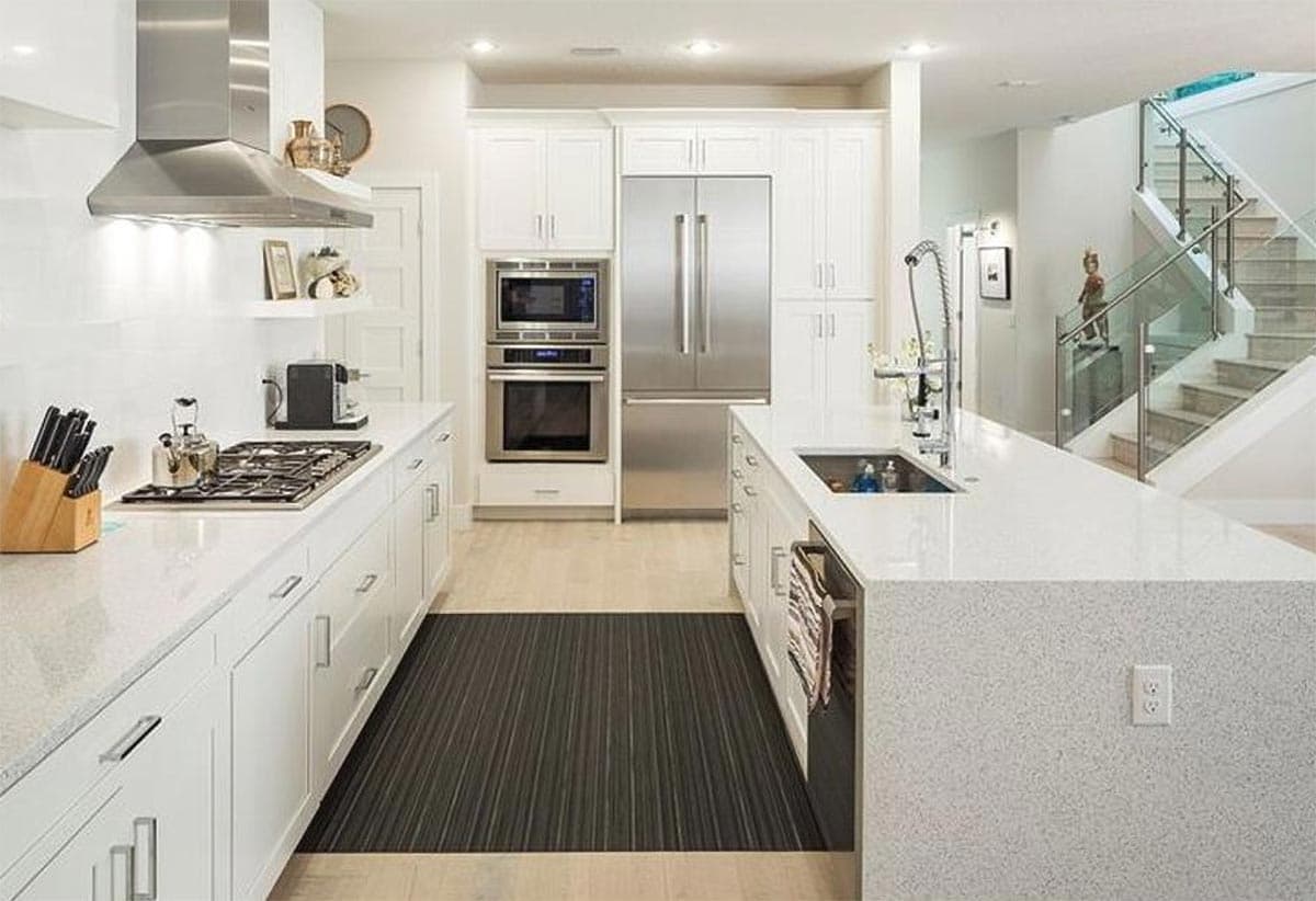 Modern kitchen with white cabinets, stainless steel appliances, quartz countertops, and island. Stairs with glass railing visible.