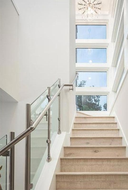 Staircase with glass railings, wood treads, and a vertical bank of windows topped by a chandelier.