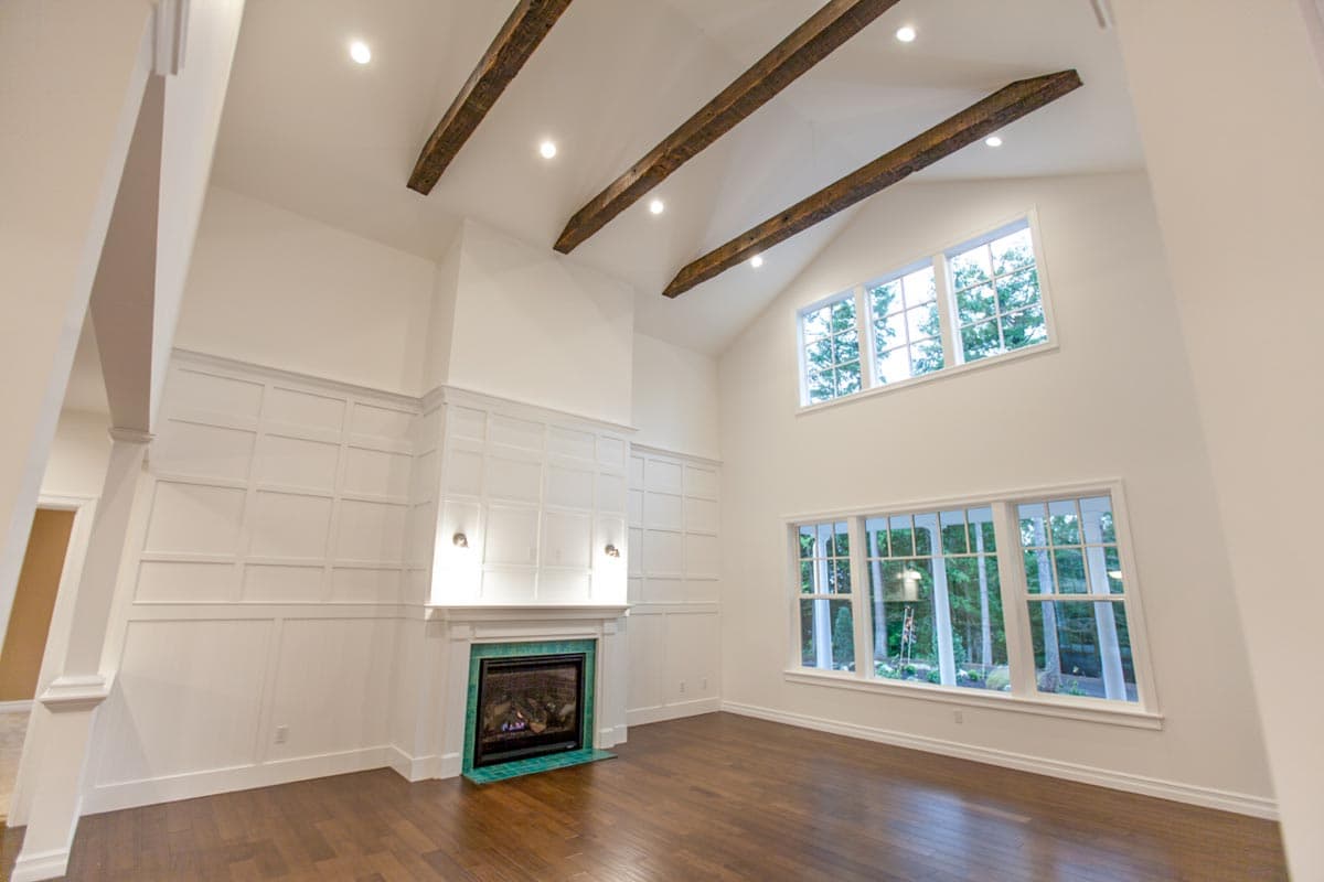 Interior view of a great room with a fireplace, wainscoting, vaulted ceiling with exposed beams, and large windows.
