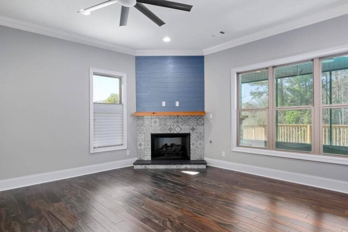 Living room with corner fireplace featuring patterned tile surround, shiplap accent, and wooden mantel. Large windows overlook deck.