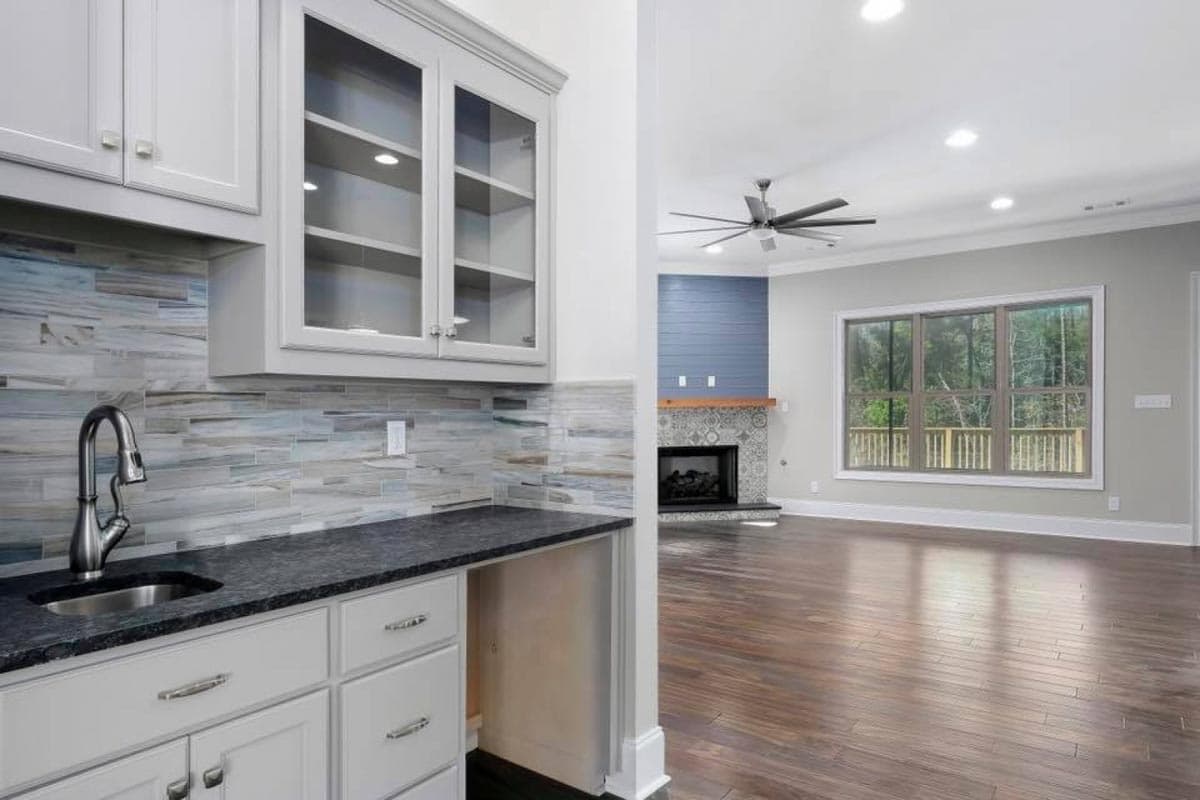 Wet bar with glass-front cabinets and dark countertop, adjacent to a great room with fireplace and large window.