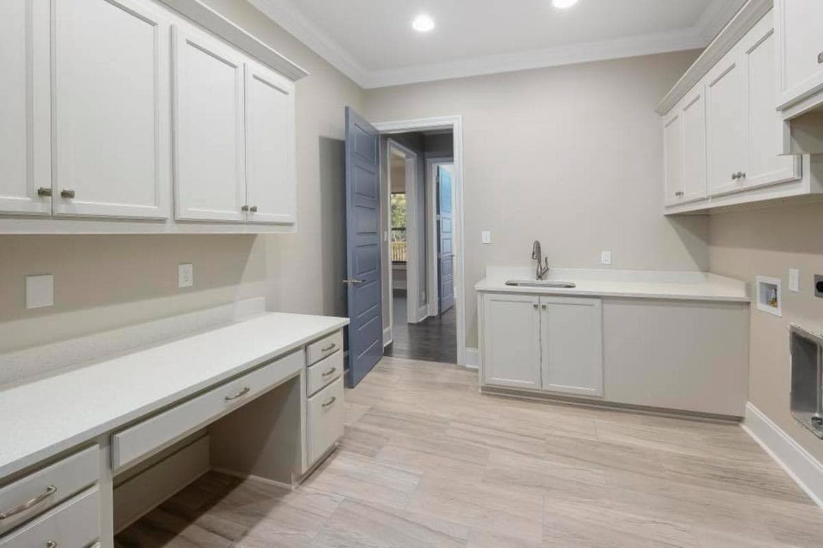 Interior view of a laundry room with white cabinetry, a built-in desk, sink, and visible doorway to another room.