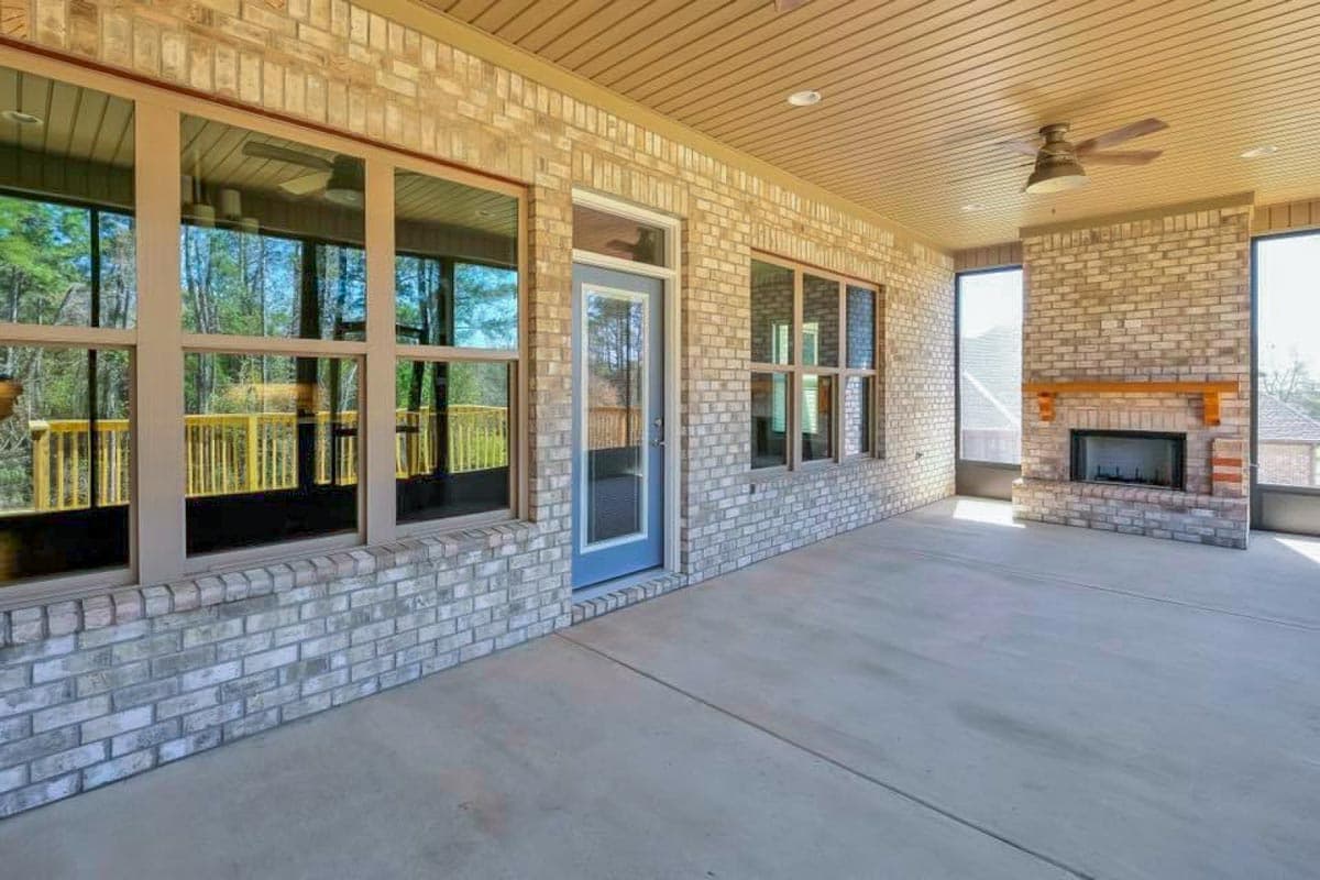Covered porch with brick exterior, fireplace, ceiling fan, and large windows overlooking a wooden deck.