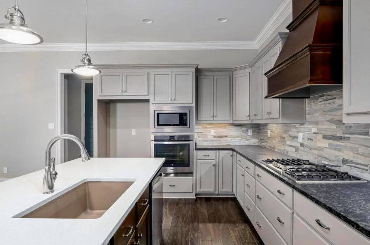 Kitchen with light gray cabinetry, stainless steel appliances, a large island with sink, and a gas range with a dark wood hood.