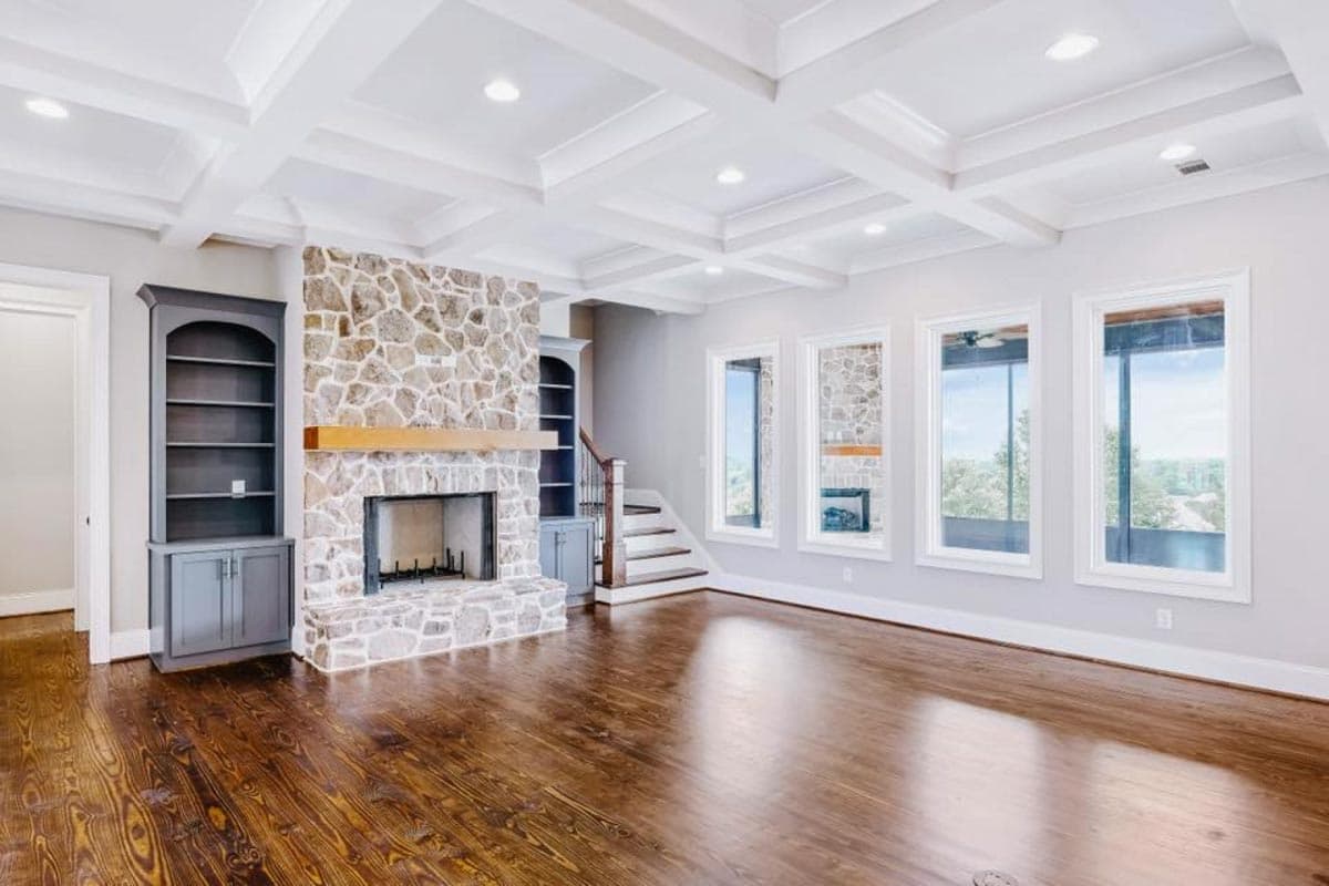 Living room with coffered ceiling, stone fireplace, built-in bookcases, and large windows.