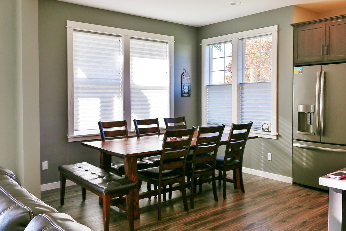 Dining area with wooden table, chairs, bench, and stainless steel refrigerator next to windows.