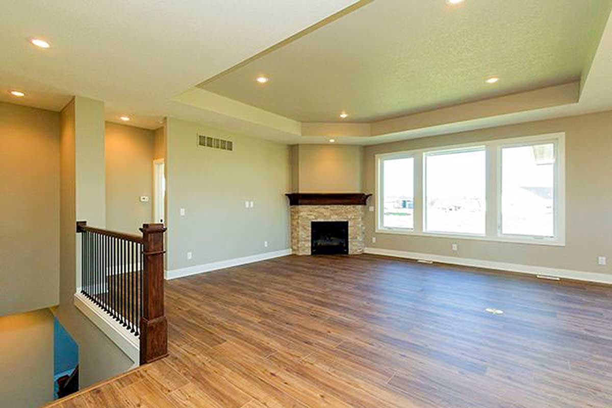 Living room with stone fireplace, tray ceiling, large windows, and wood flooring. Stair railing visible.