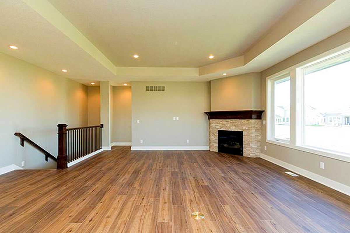 Interior view of a great room with a stone fireplace, large windows, tiered ceiling, and wood flooring leading to stairs.
