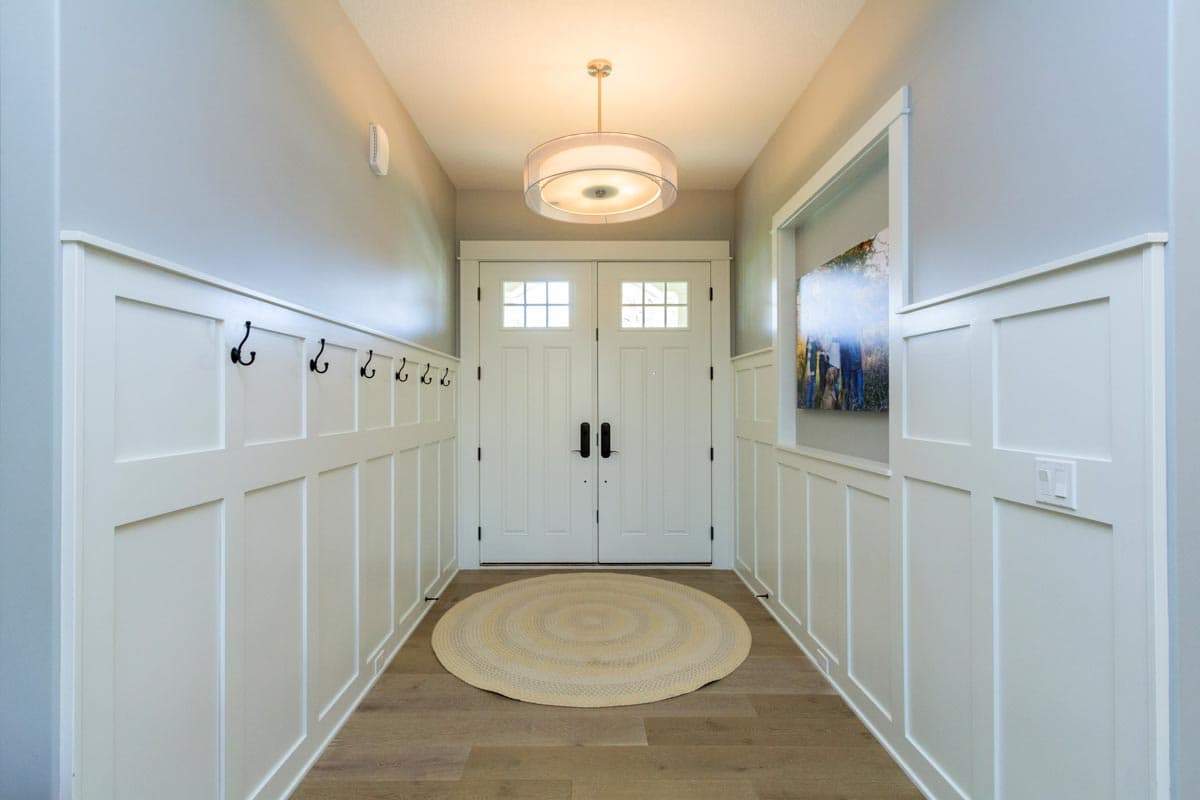 Interior view of a foyer with paneled walls, coat hooks, double doors with glass panes, and a circular rug.