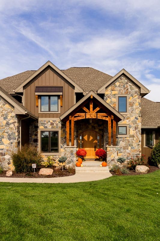 House plan exterior: stone and wood siding, prominent timber-framed gable entry, multi-level gables, and pitched roof.