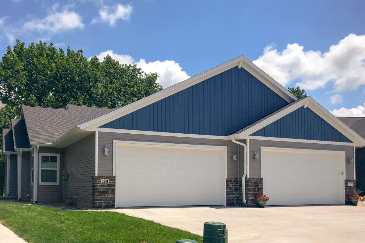 House plan exterior with two-car garage doors, grey horizontal siding, and blue vertical gables.