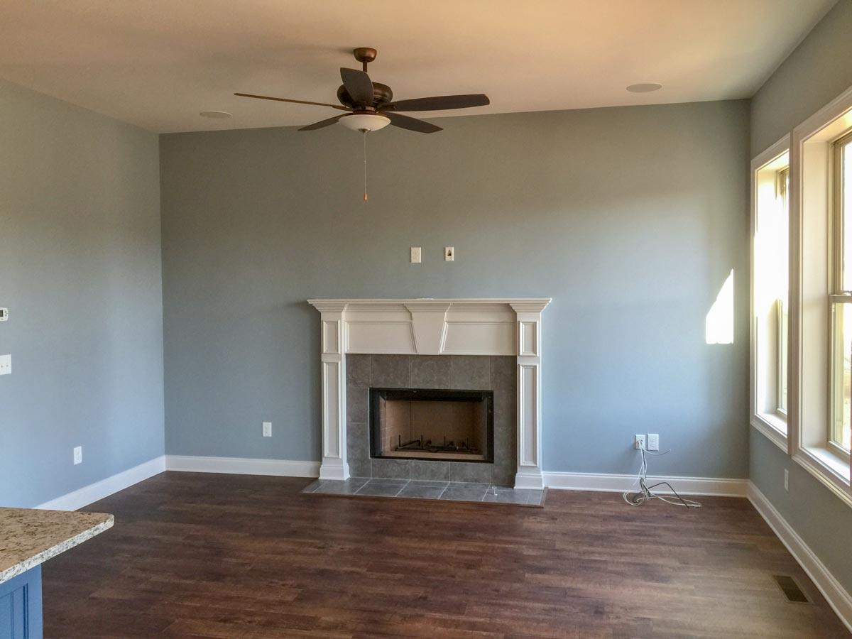 Interior view of a living room featuring a tiled fireplace with ornate mantel, ceiling fan, and large windows.