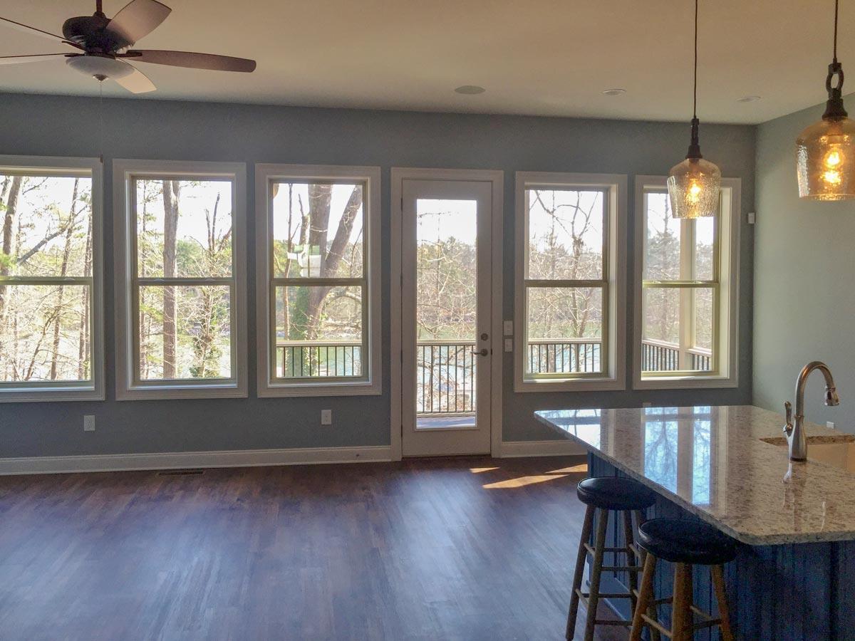 Interior view of a home with a large kitchen island, pendant lights, and multiple windows overlooking a wooded area and deck.