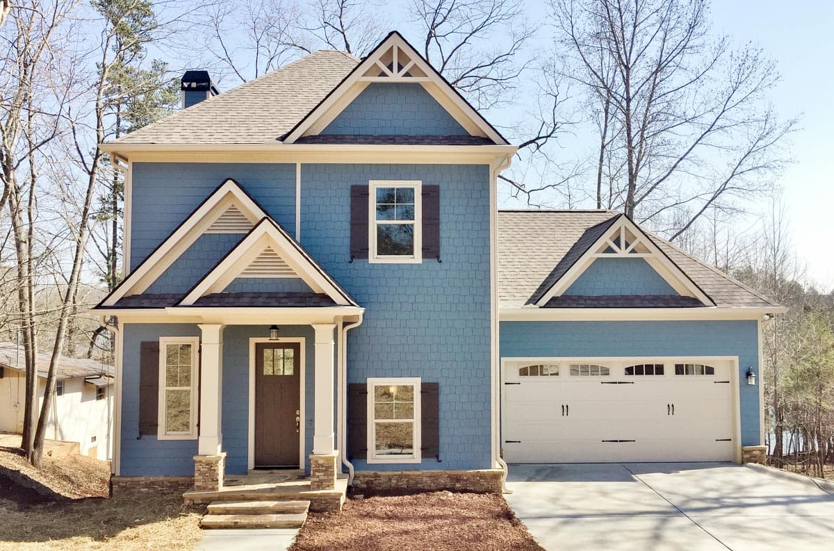 House plan exterior: two-story blue home with gables, decorative trim, covered front porch with stone pillars, and a two-car side-entry garage.
