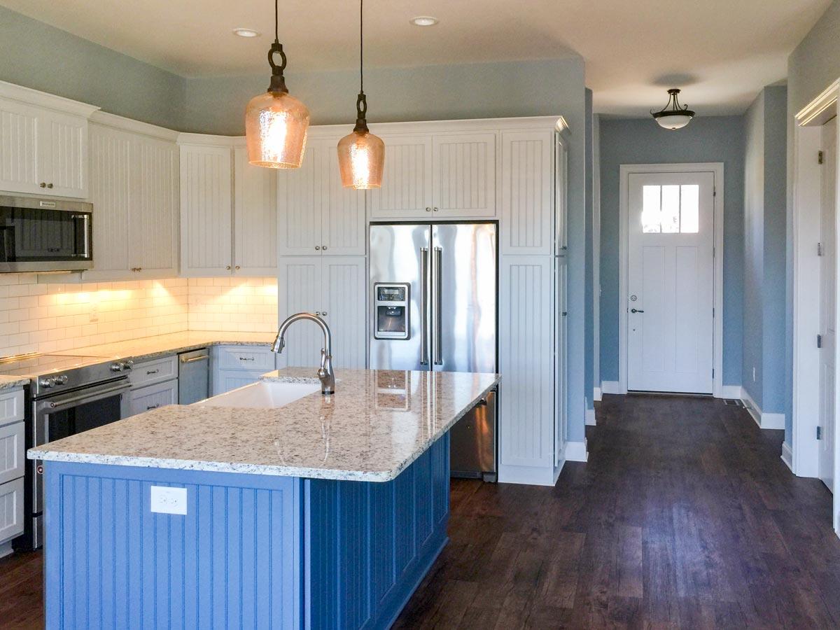 Kitchen with white beadboard cabinets, granite island, and stainless steel appliances leading to a hallway with a white front door.