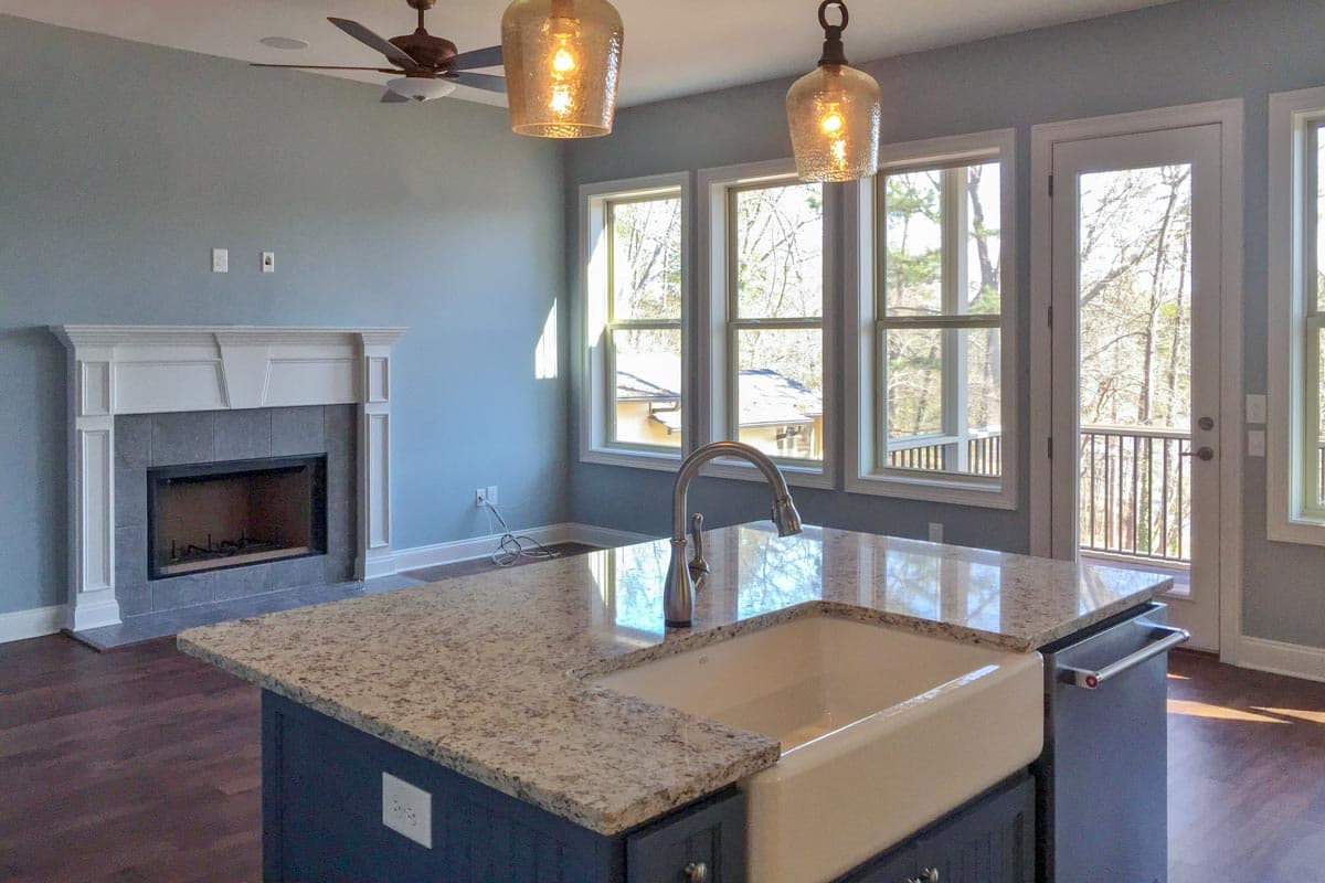 Kitchen island with farmhouse sink, fireplace with tile surround, ceiling fan, and windows overlooking a wooded area.