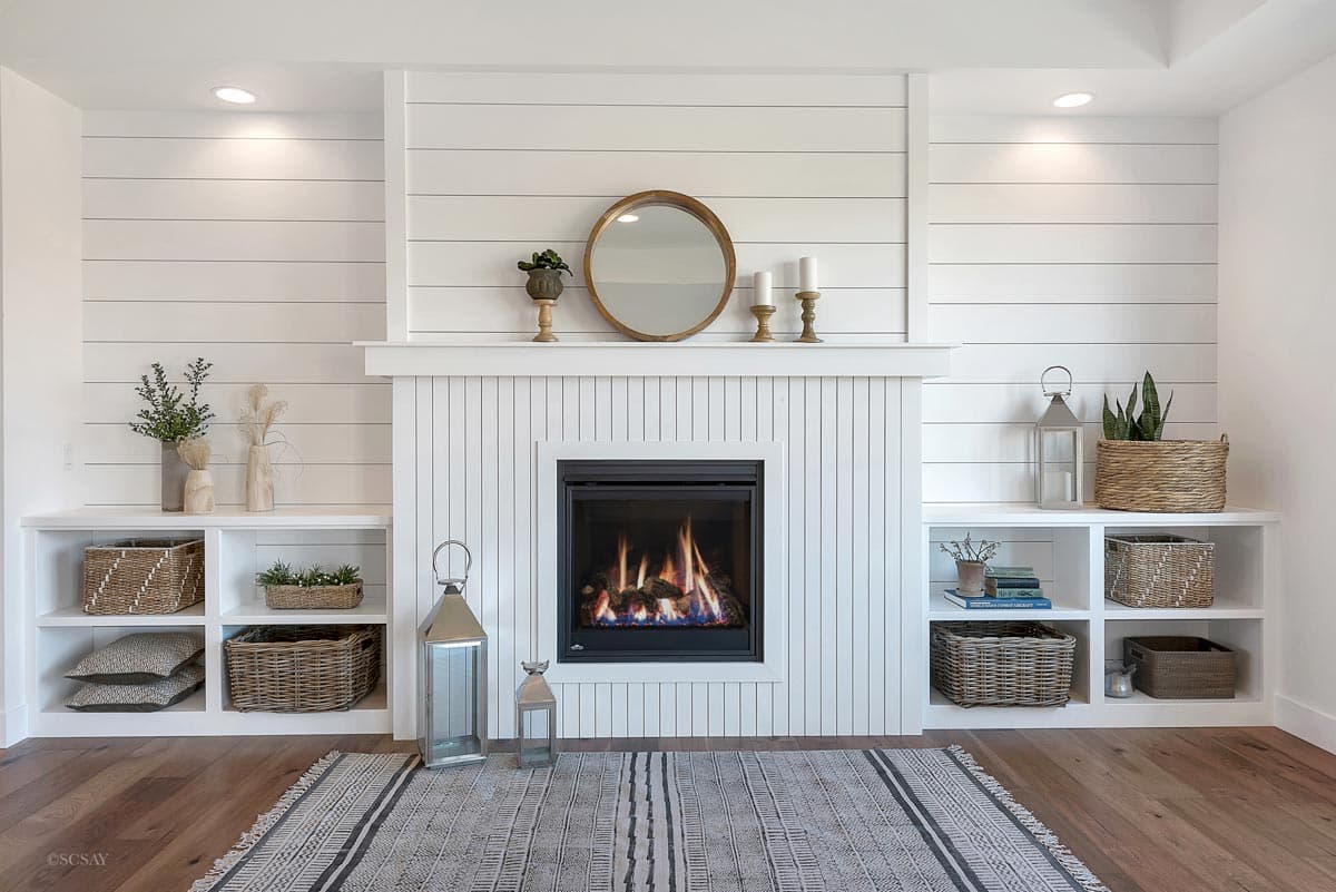 Living room fireplace with shiplap and beadboard surround, flanked by built-in shelves with baskets and decor.