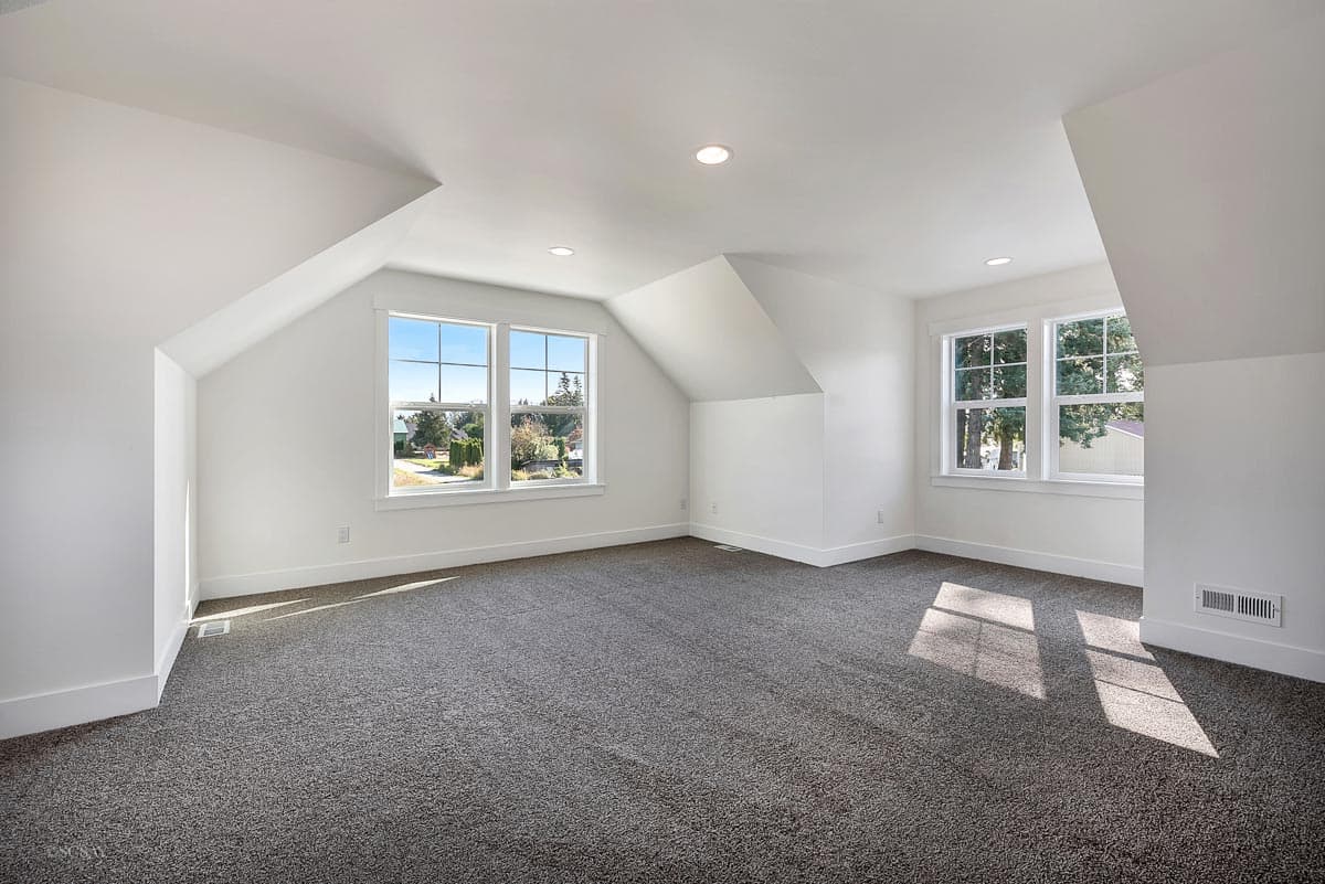 Interior view of an unfinished room with sloped ceilings, multiple windows, and carpeted floor.