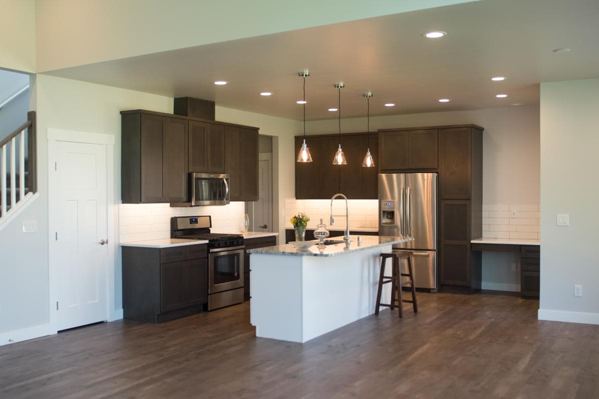 Modern kitchen with dark cabinets, granite island with pendant lights, stainless steel appliances, subway tile backsplash, and adjacent stairs.