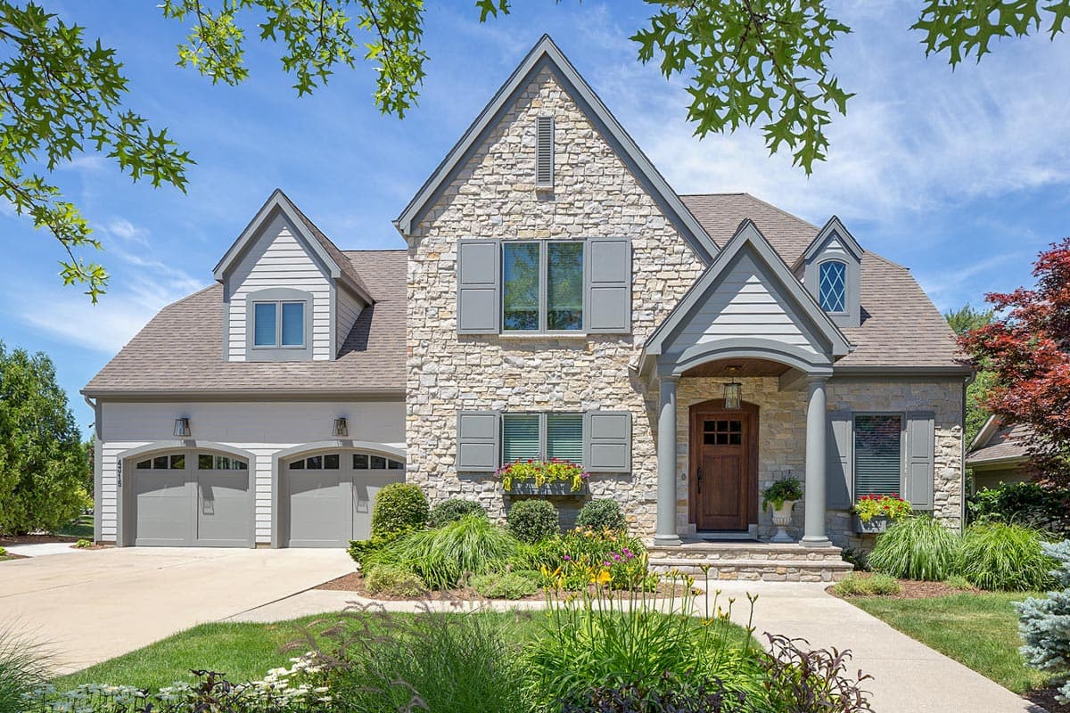 House plan exterior. Two-story with stone facade, gabled roof, and arched garage doors. Features a covered porch and dormers.