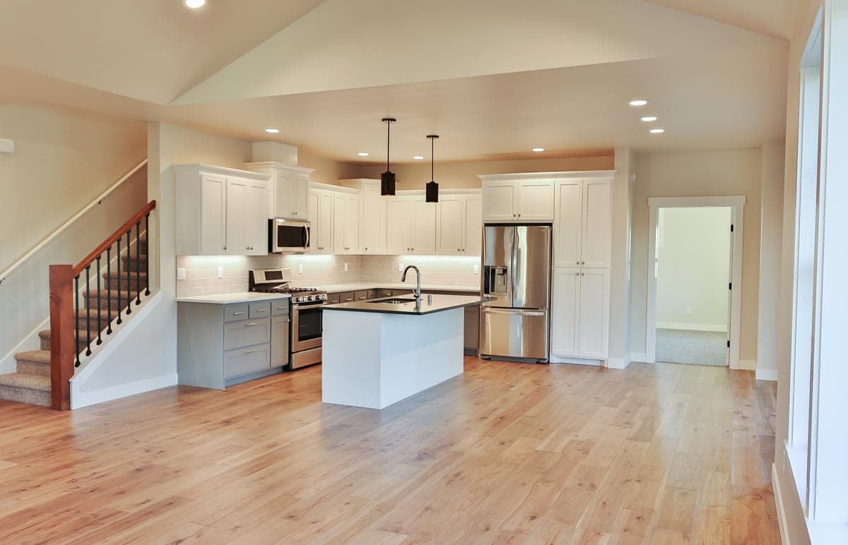 Open concept kitchen with white cabinets, island, stainless steel appliances, and vaulted ceiling. Stairs to upper level visible.