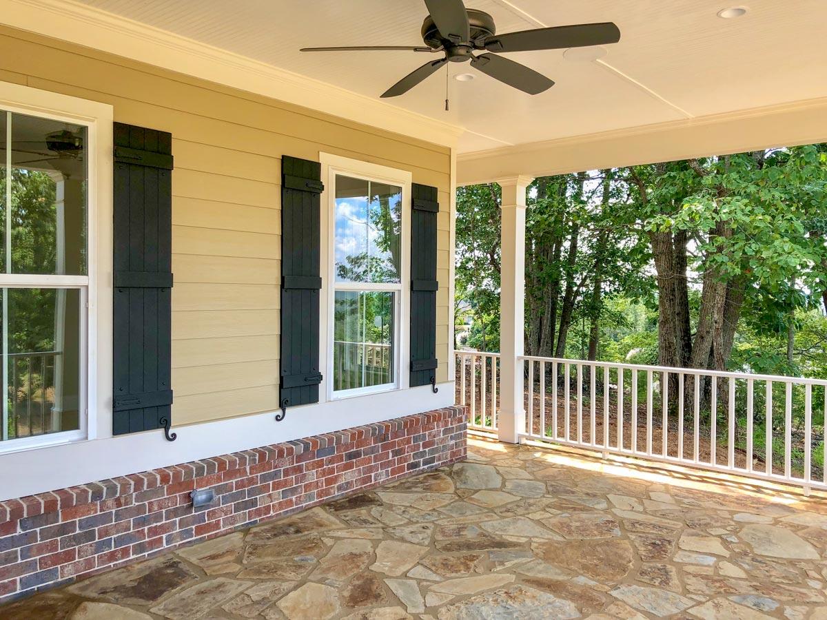 House plan exterior: Covered porch with stone patio, white railings, windows with black shutters, and brick foundation.