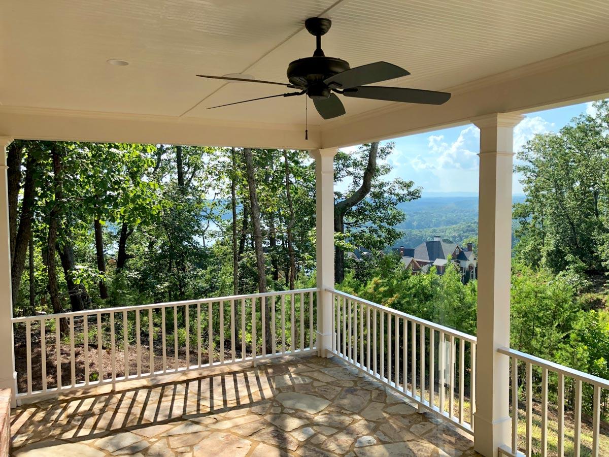 Covered porch with stone floor, white railings, and ceiling fan, overlooking wooded landscape and distant homes.