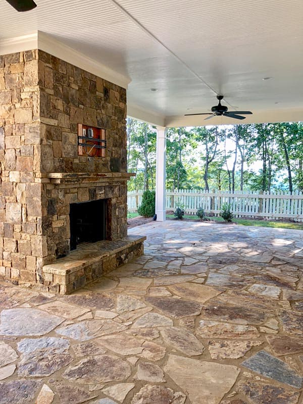 Covered patio with a large stone fireplace, stone patio flooring, and ceiling fan.