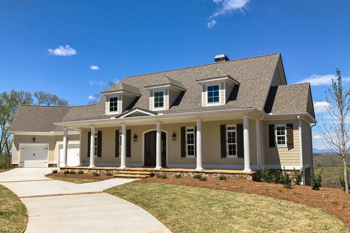 House plan exterior. Two-story home with a wrap-around porch, dormers, and a gabled roof. Attached garage is oriented to the side.