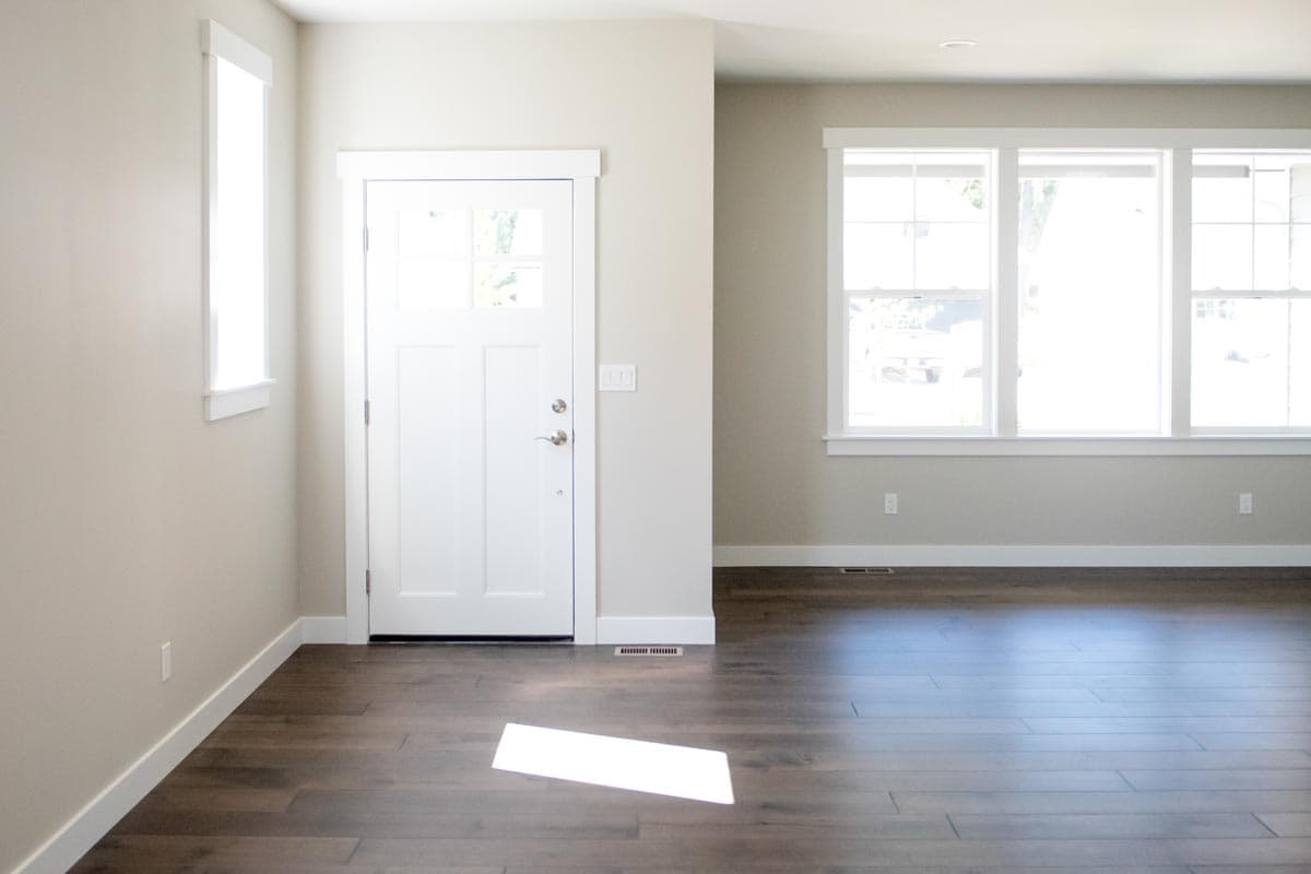 Interior view of a house entrance with a white door and a wide window featuring three panes. Dark wood floors.