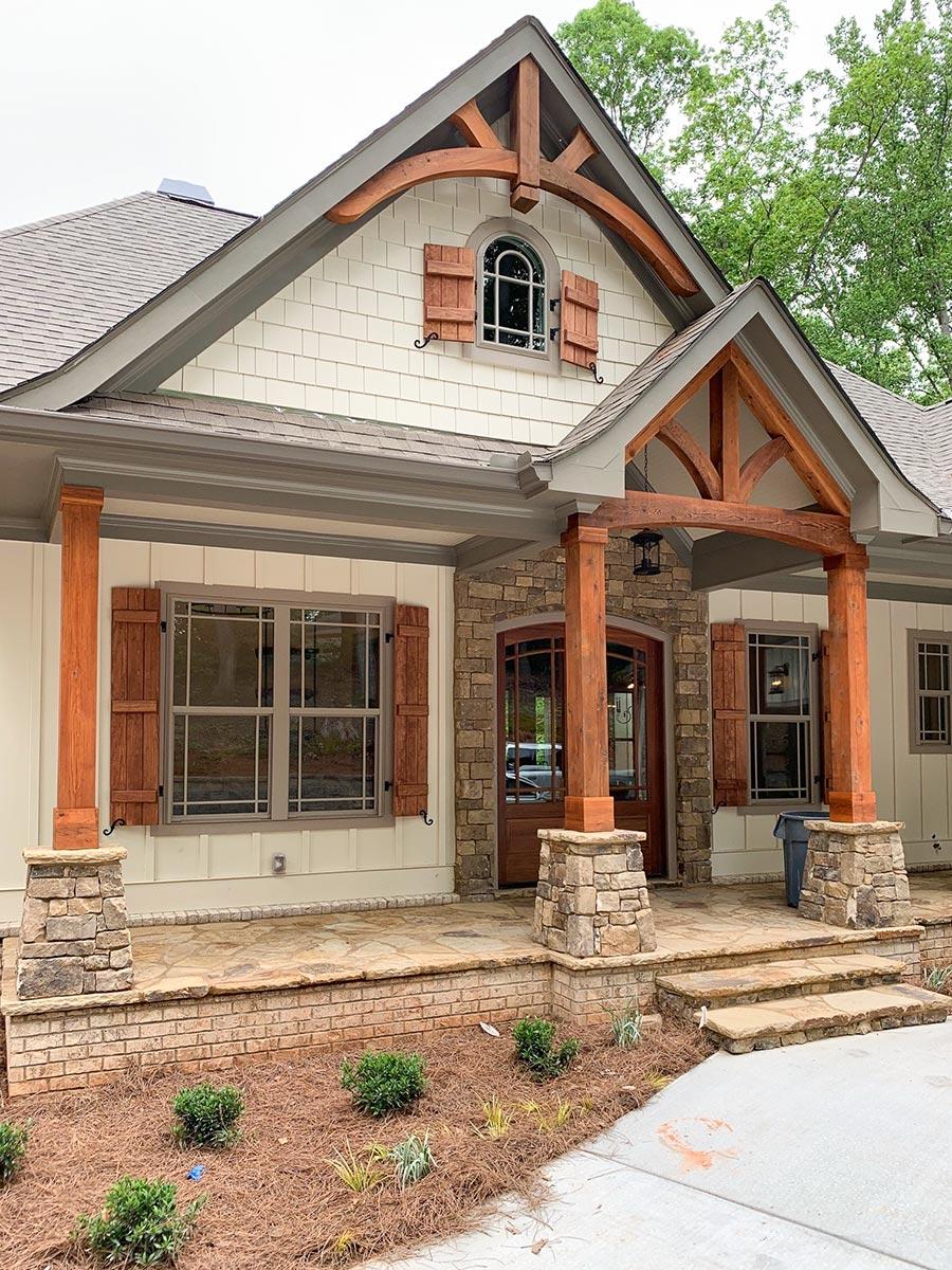 House plan exterior with prominent wooden beams, stone accents, arched window with shutters, and covered porch.