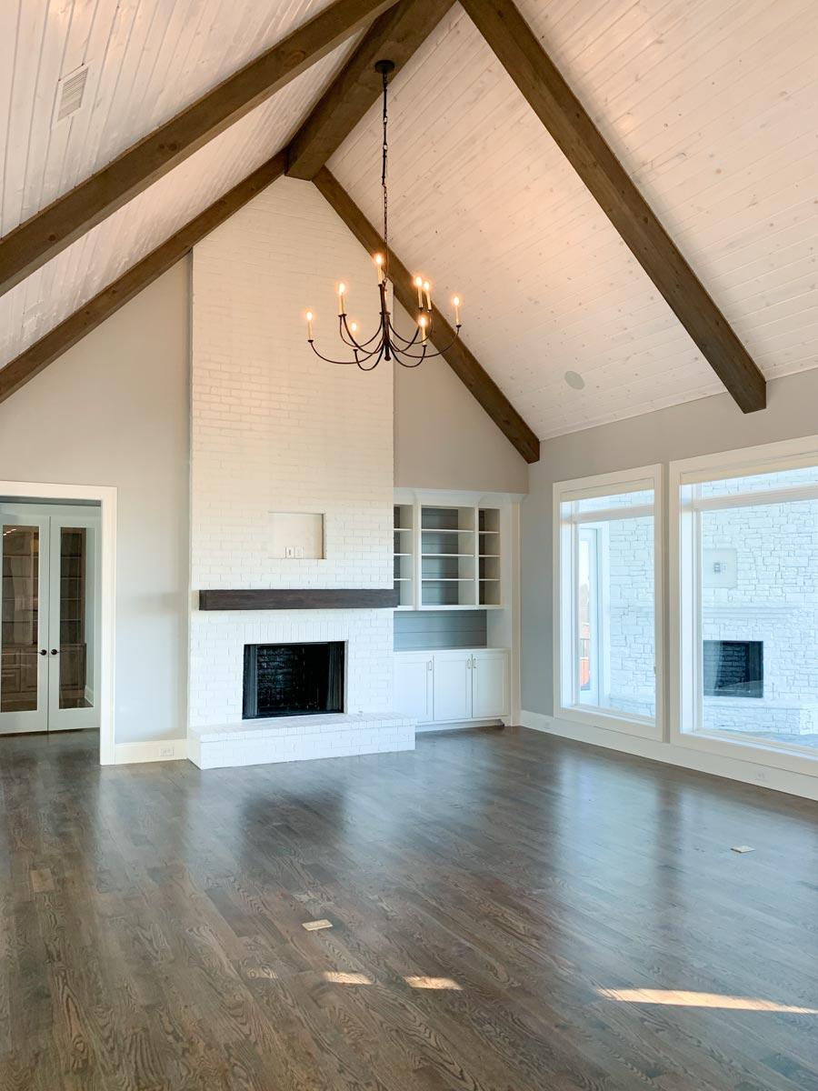 Great room with vaulted ceiling, exposed beams, white brick fireplace, built-in shelving, and large windows.