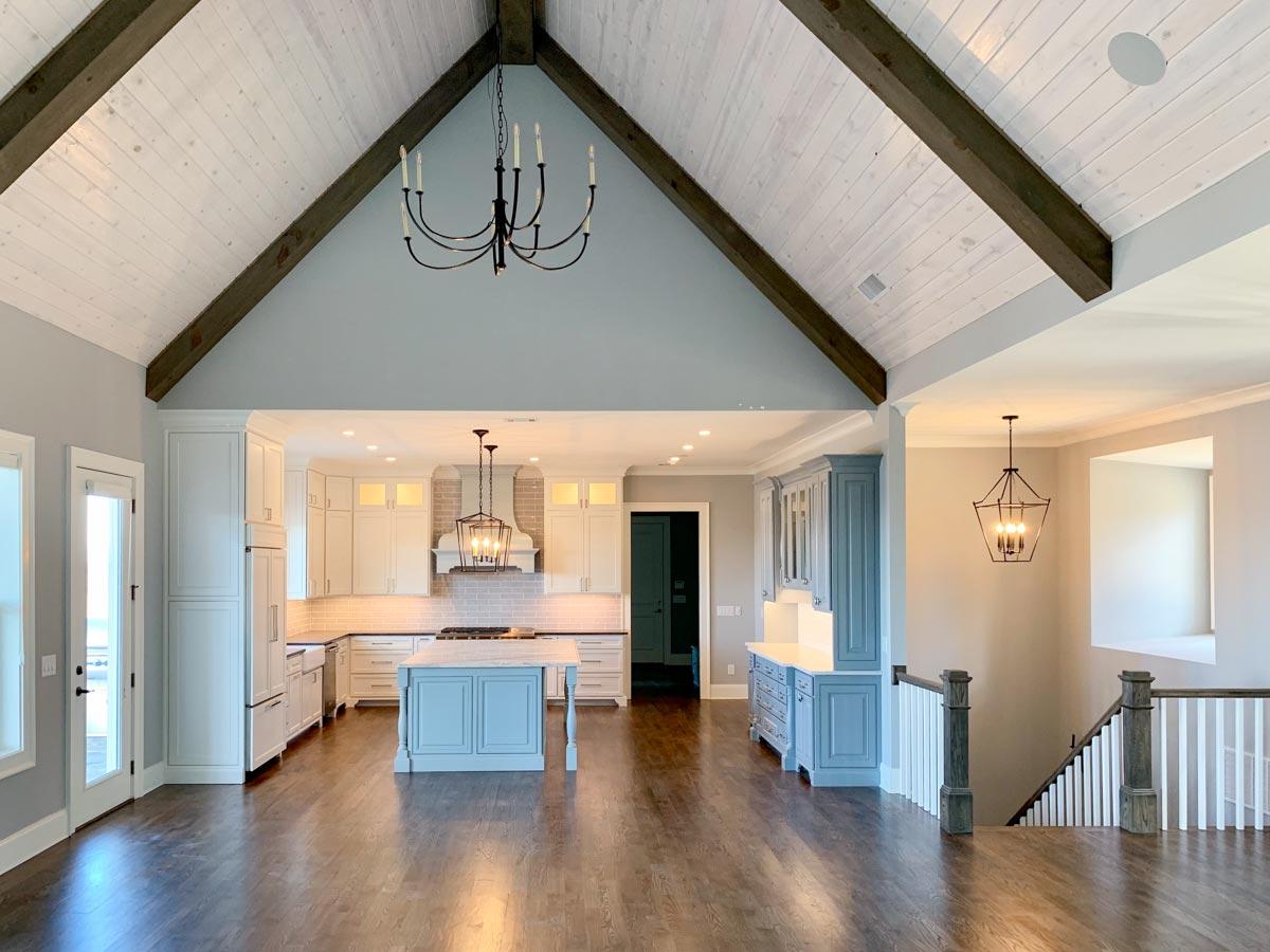Open concept living space with vaulted wood-beamed ceiling, kitchen with island and white cabinetry, and a staircase.
