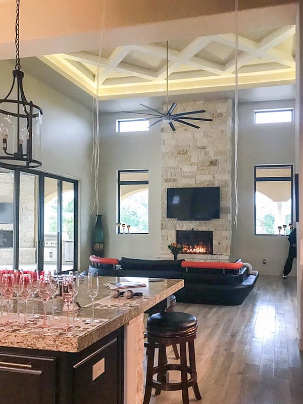 Interior view of great room with stone fireplace, coffered ceiling with lighting, large ceiling fan, and kitchen island.